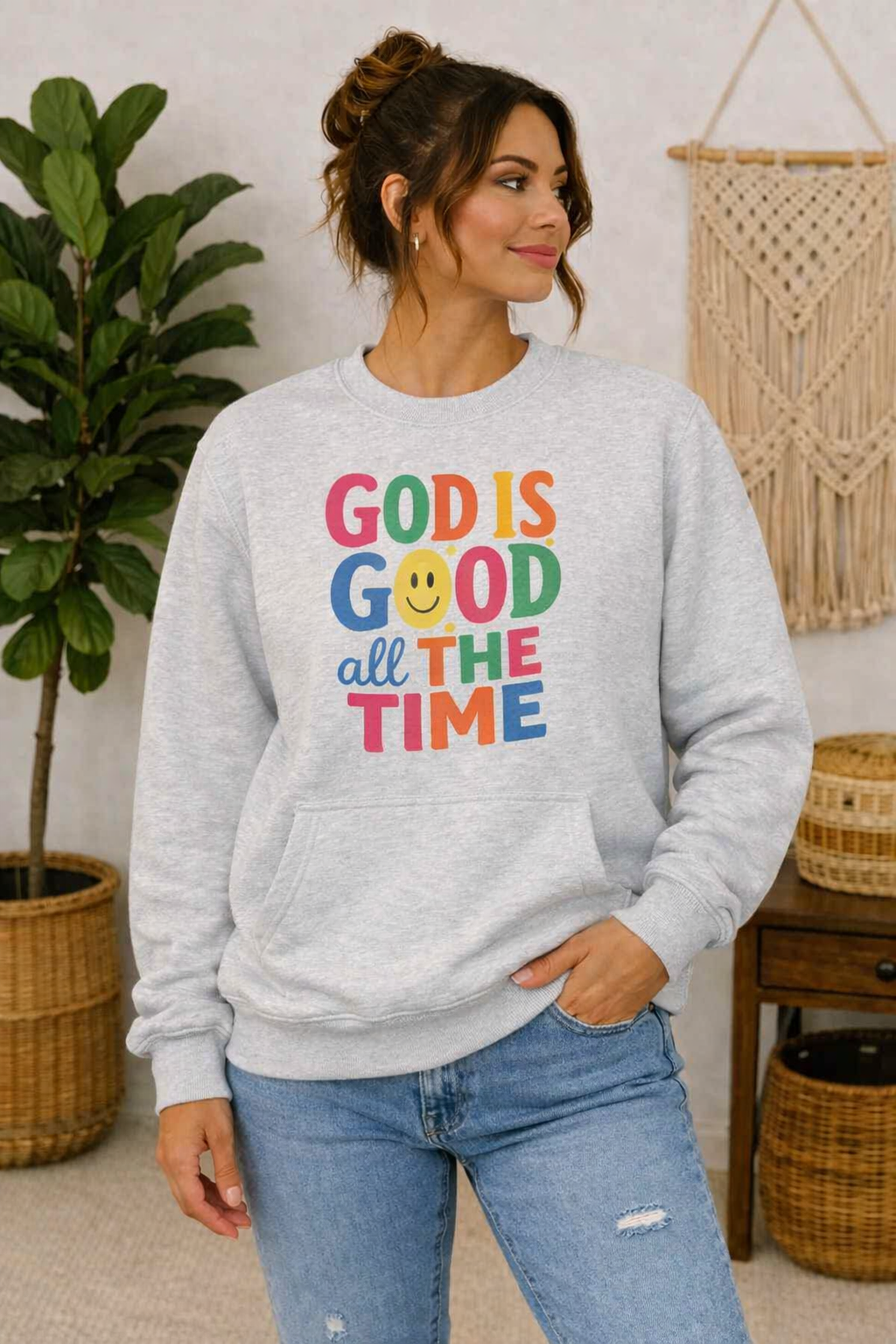A woman wearing a light gray crewneck sweatshirt with a colorful graphic that reads 'GOD IS GOOD all THE TIME' with a smiley face, paired with blue jeans in a cozy indoor setting.