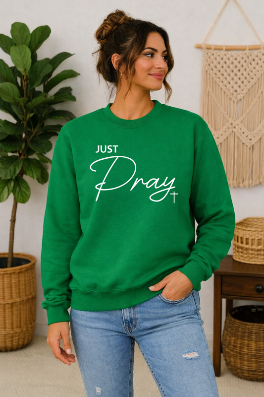 A woman wearing a bright green crewneck sweatshirt with the text 'JUST Pray' in white script, paired with light blue jeans, standing in a decorated room with a plant and macrame wall hanging.
