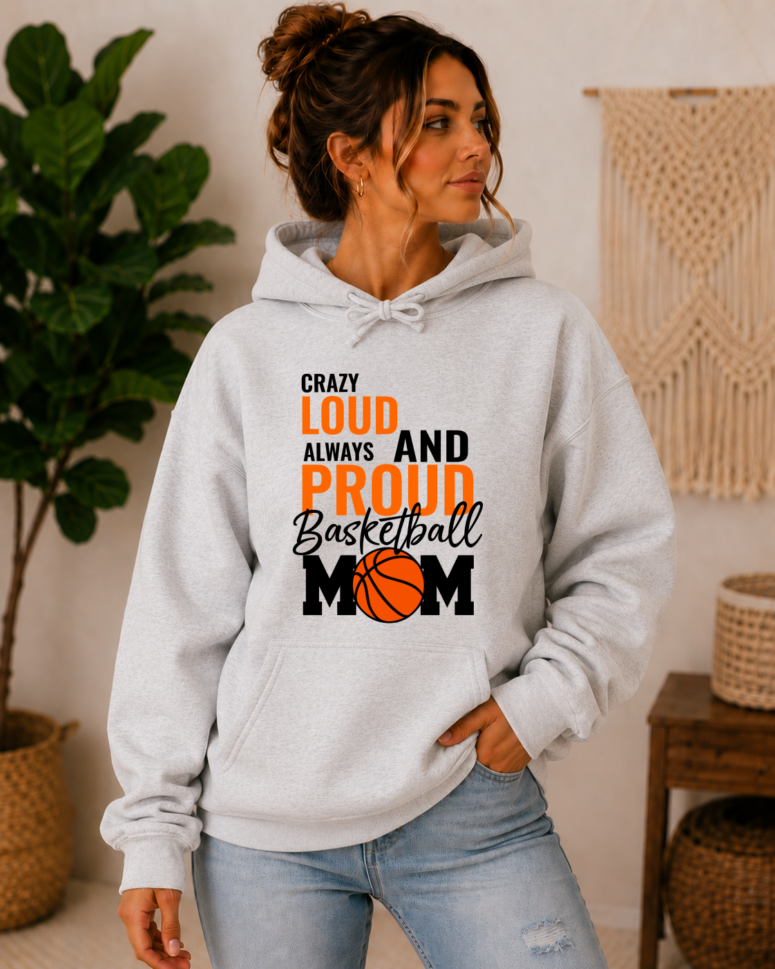 A woman wearing a light gray pullover hoodie with a graphic that reads 'Crazy Loud Always and Proud Basketball Mom' in black and orange lettering, paired with light blue jeans in a cozy indoor setting.