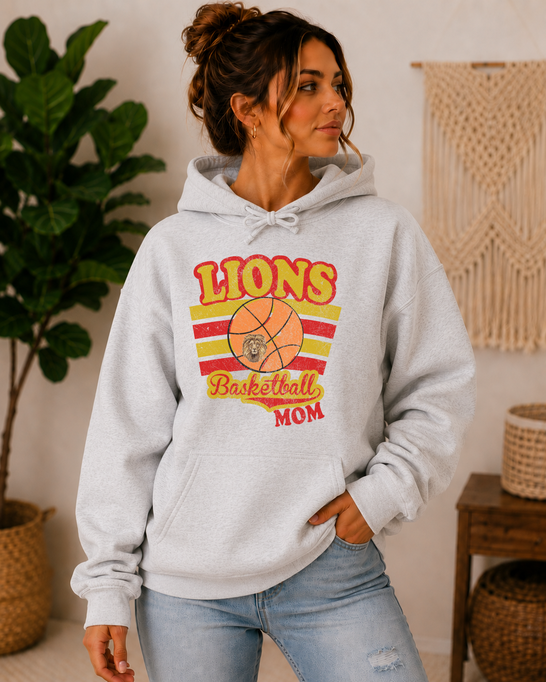 A woman wearing a light gray heather hoodie with a retro-style 'Lions Basketball Mom' graphic on the front, paired with light-wash jeans in a cozy indoor setting.
