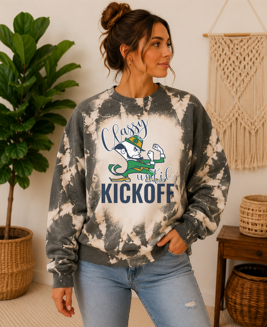 A woman wearing a gray and white tie-dye crewneck sweatshirt with a St. Patrick's Day graphic that reads 'Happy until KICKOFF' with a cartoon leprechaun, paired with light blue jeans in a cozy indoor setting.