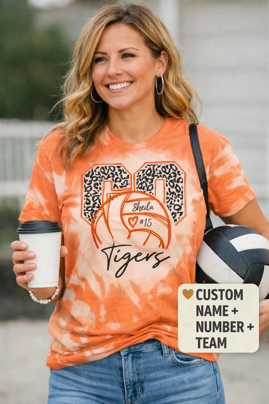 A woman wearing an orange tie-dye t-shirt with a custom basketball graphic that reads 'Shelton #15 Tigers' with leopard print details. She is holding a coffee cup and a volleyball, with a text overlay on the bottom right that says 'CUSTOM NAME + NUMBER + TEAM'.