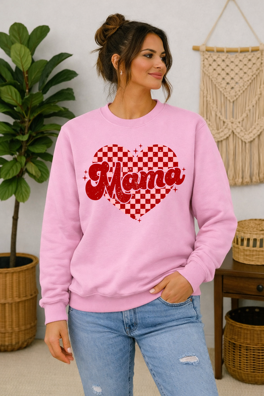 A woman wearing a light pink crewneck sweatshirt with a red and white checkered heart graphic and the word 'Mama' in red script, paired with light blue jeans, standing in a cozy room with a plant and macrame wall hanging.