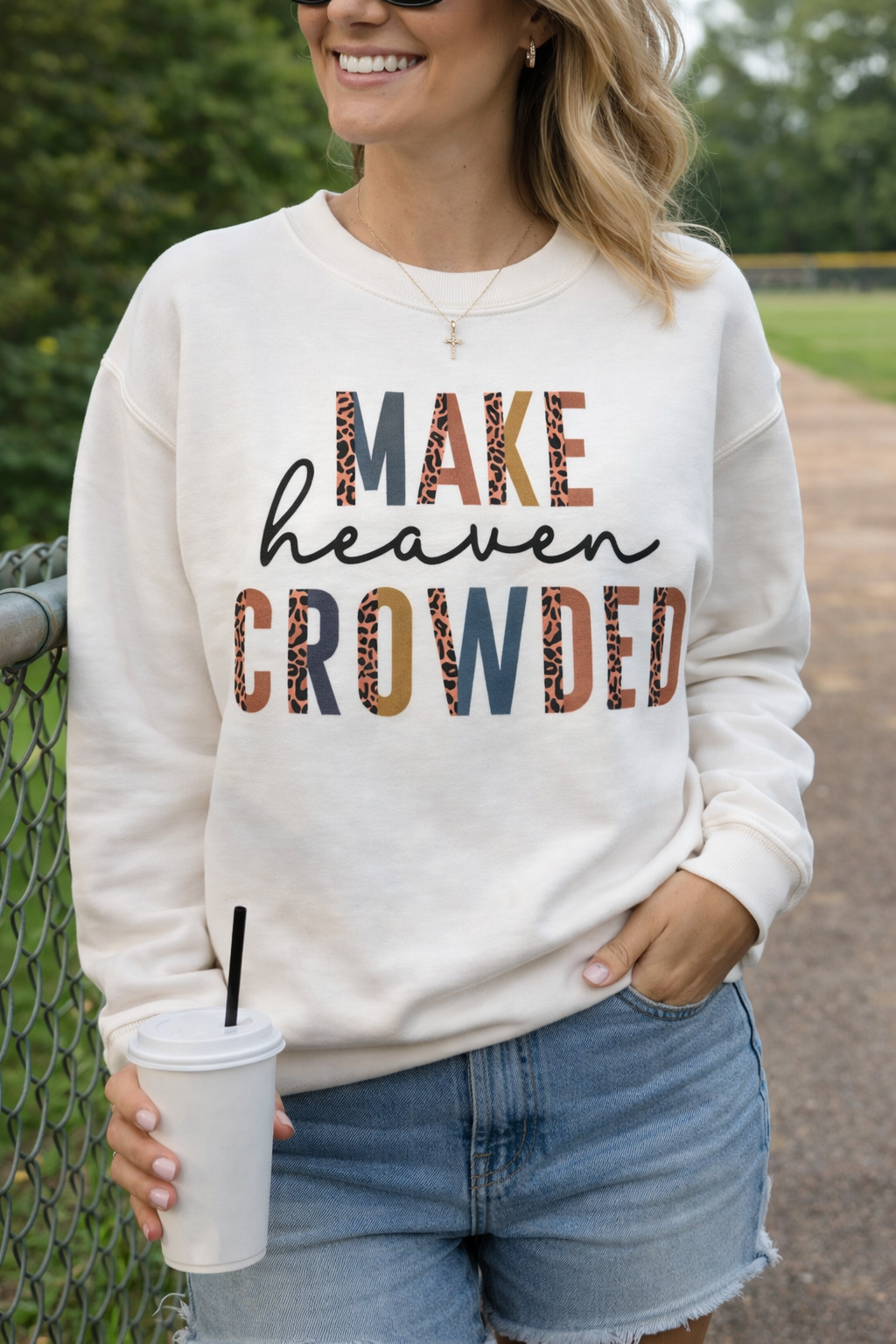 Person wearing a sweatshirt with 'MAKE heaven CROWDED' text, holding a cup outdoors.
